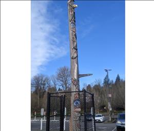 A tall wooden pole with faces and shapes carved into it. The pole is surrounded by black chain link fencing. The pole is in the middle of a parking lot with forest and blue sky in the background. 